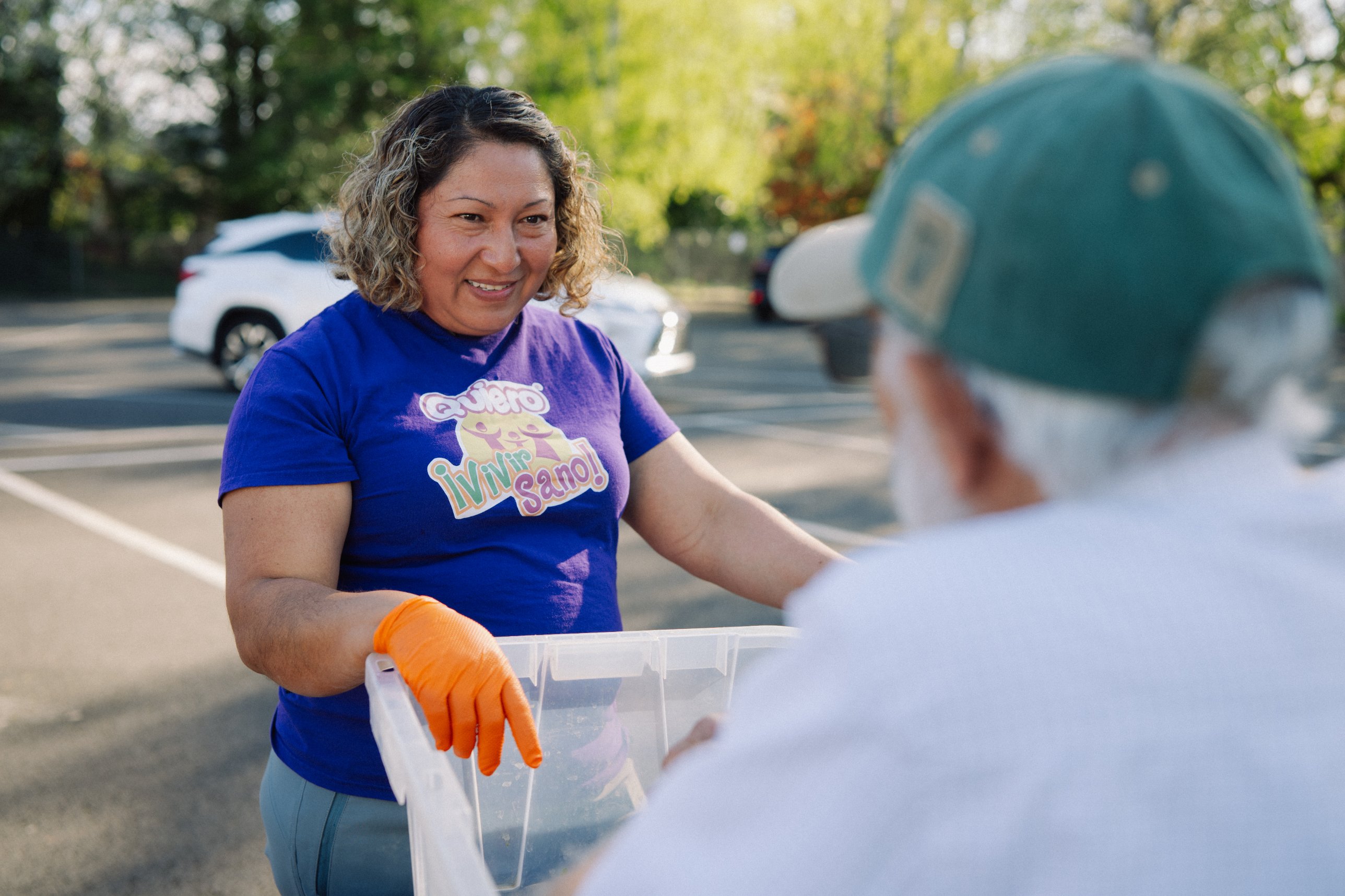 Volunteer in a purple shirt and orange glove smiling at a neighbor over a food crate at an outdoor distribution